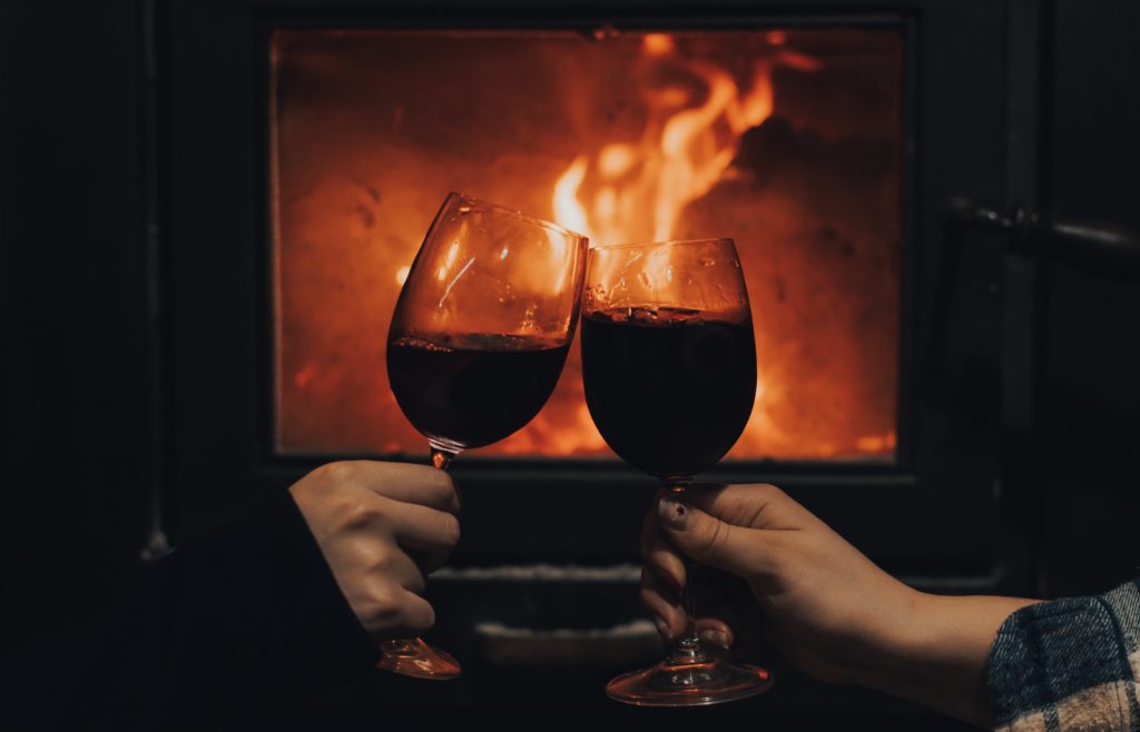 Two people in front of a fireplace each holding a glass of Malbec red wine and raising their glasses to cheers each other.